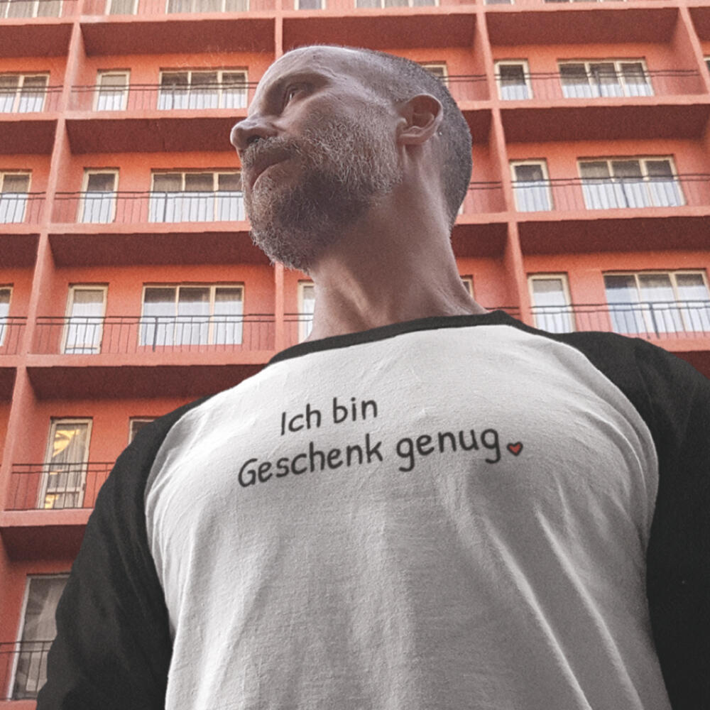 Foto eines ernst blickenden Mannes, der ein Baseballshirt trägt mit dem Aufdruck „Ich bin Geschenk genug“ mit kleinem roten Herz und darunter eine freundliche Strichfigur mit Weihnachtsmütze, die selbst als Geschenk eingepackt dasteht.